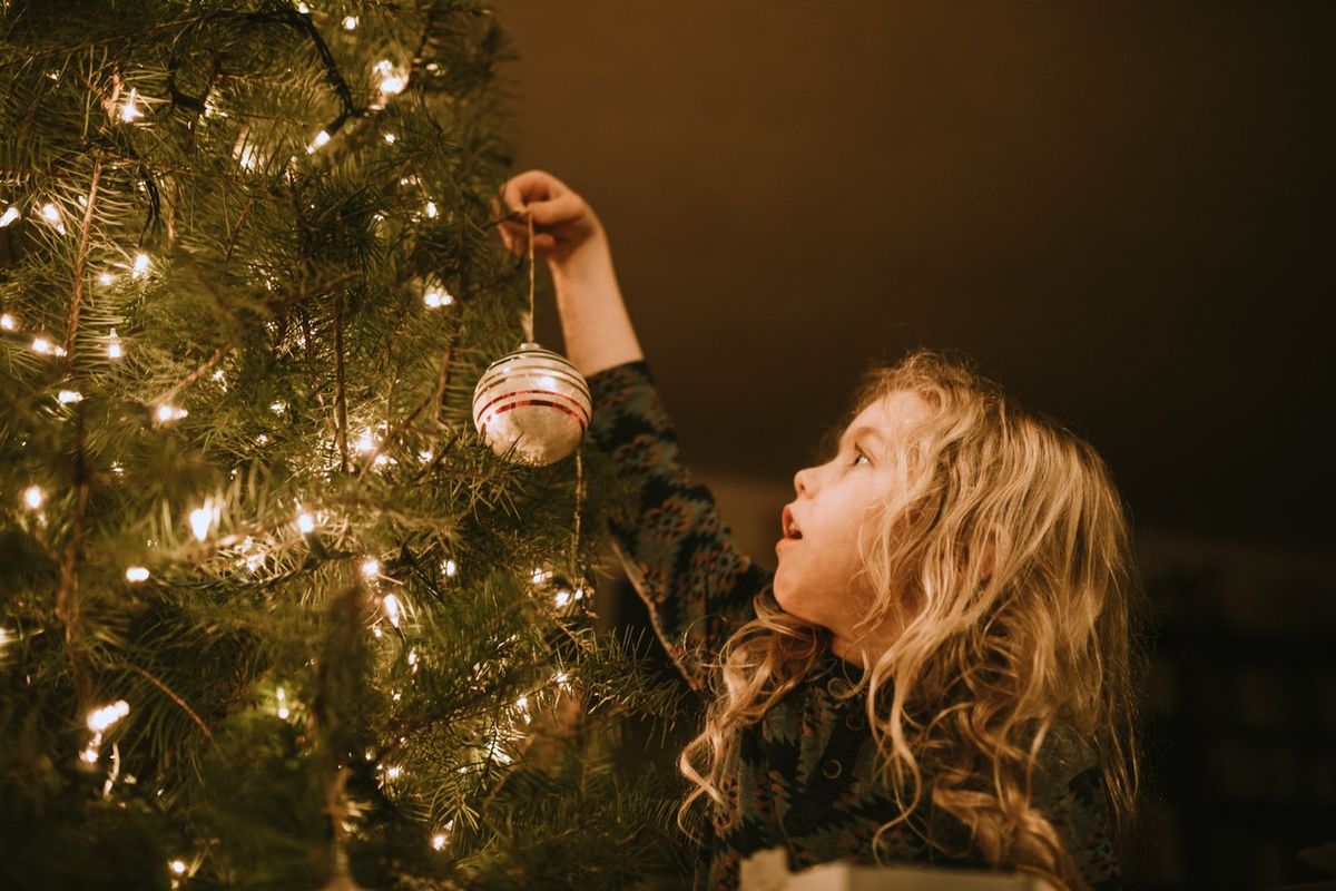 A child reaches up high to hang an ornament on the lighted Christmas tree in her home.