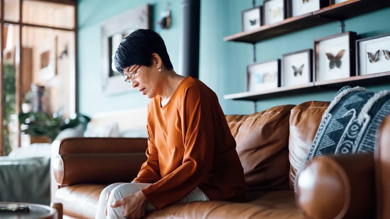 A distraught senior Asian woman feeling unwell, suffering from pain in leg while sitting on sofa in the living room at home