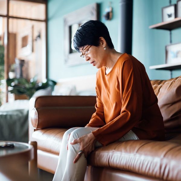 A distraught senior Asian woman feeling unwell, suffering from pain in leg while sitting on sofa in the living room at home