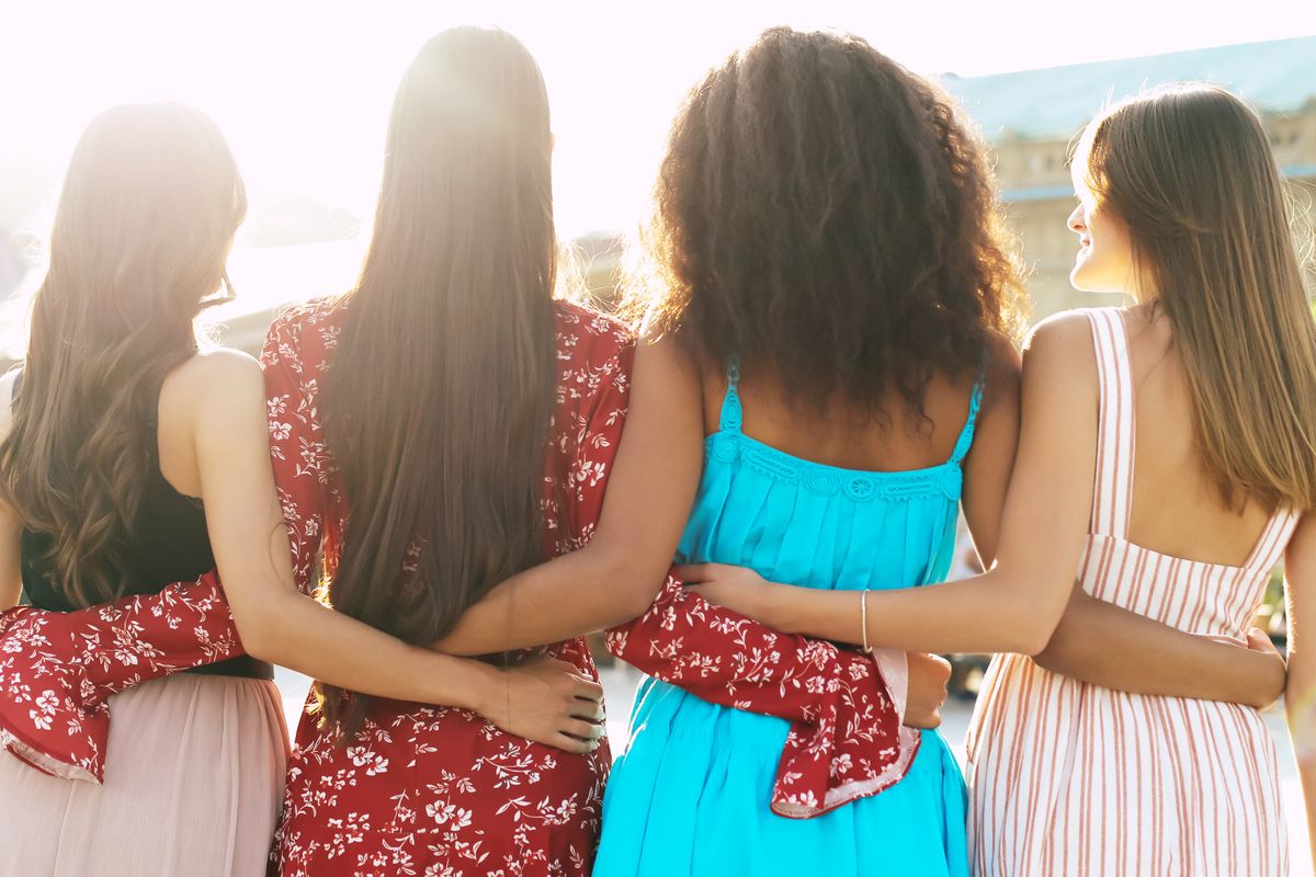 A group of four best friends in magnificent summer outfits are hugging each other and posing with their backs to the camera.