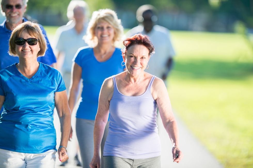 A multi-ethnic group of senior adults are exercising together by walking in the park on a sunny day.