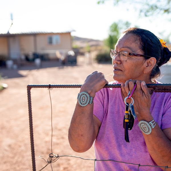 A Navajo woman standing by the gate of her home, with a faraway look in her eyes
