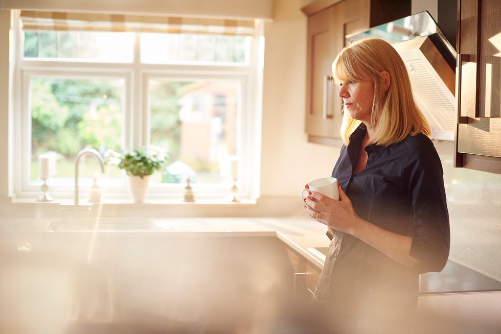 a woman stands in her kitchen and has a cuppa and a break looking to the floor in sad contemplation