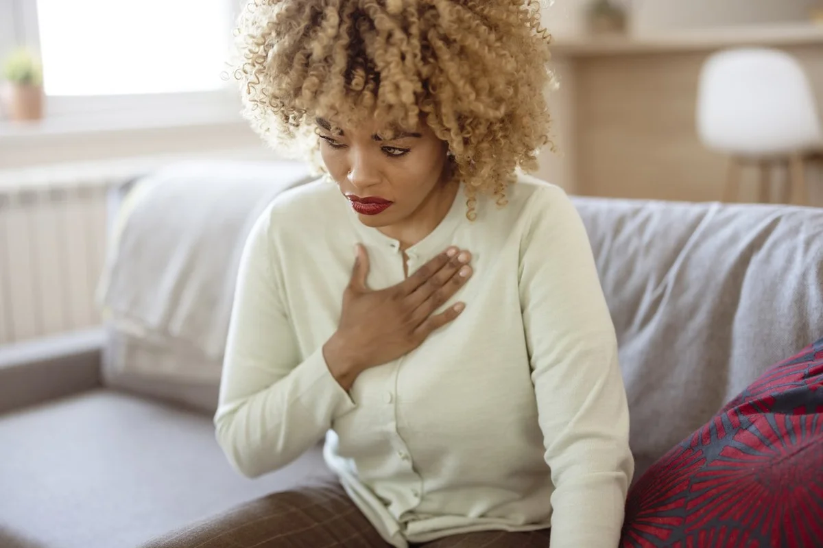 African American woman feeling sick and holding her chest in pain while sitting on the sofa in the living room