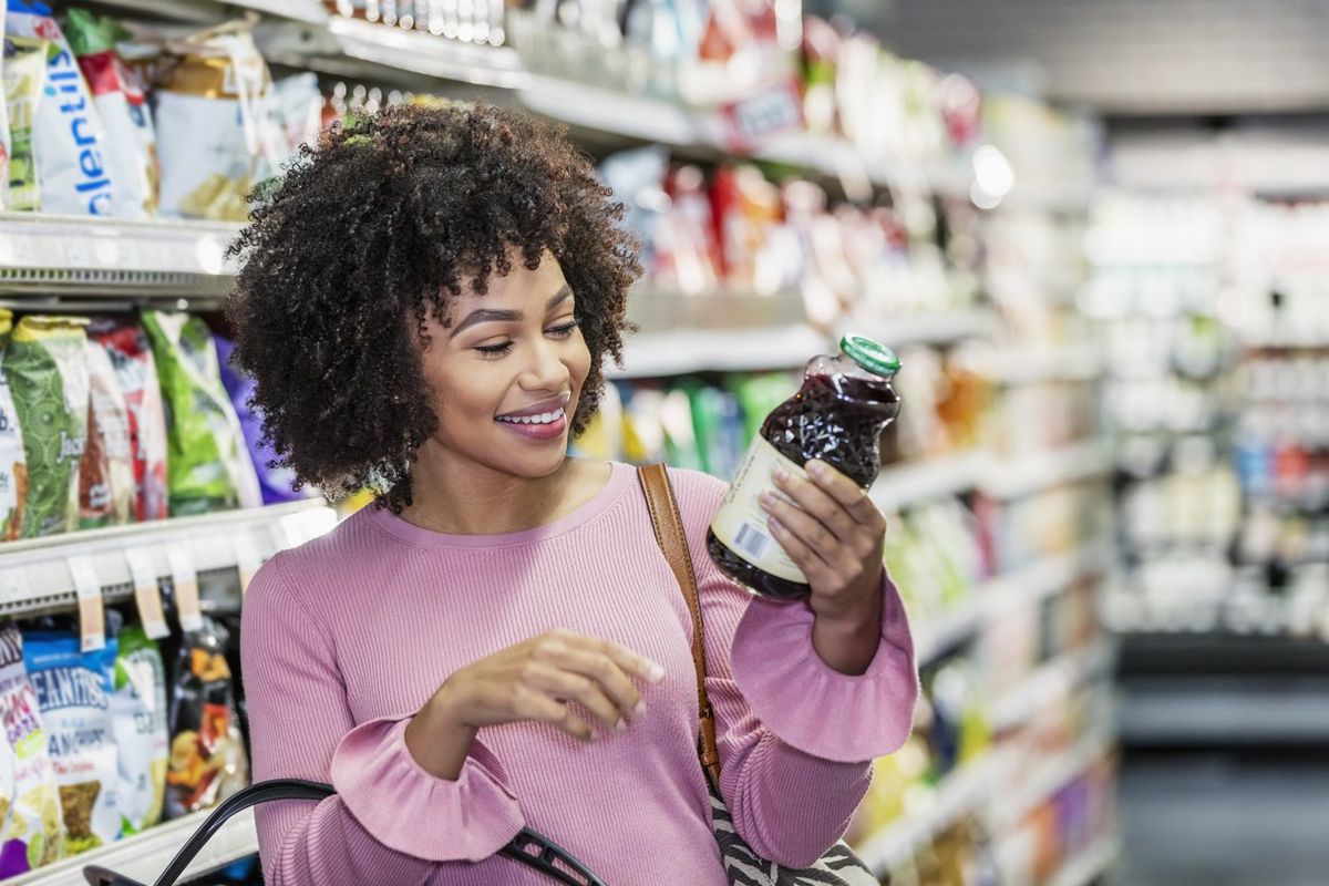 African-American woman in her 20s shopping in a grocery store, carrying a shopping basket. She is reading the ingredient label on a bottle