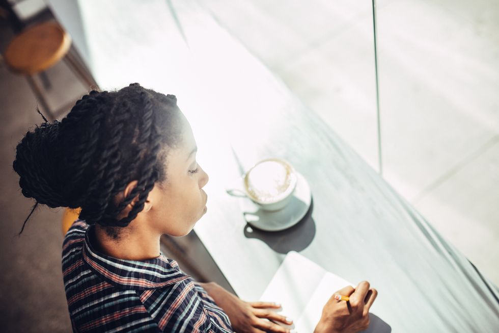 African American woman sits at the window counter of a coffee shop, enjoying a latte while writing ideas down in a small notepad journal or diary