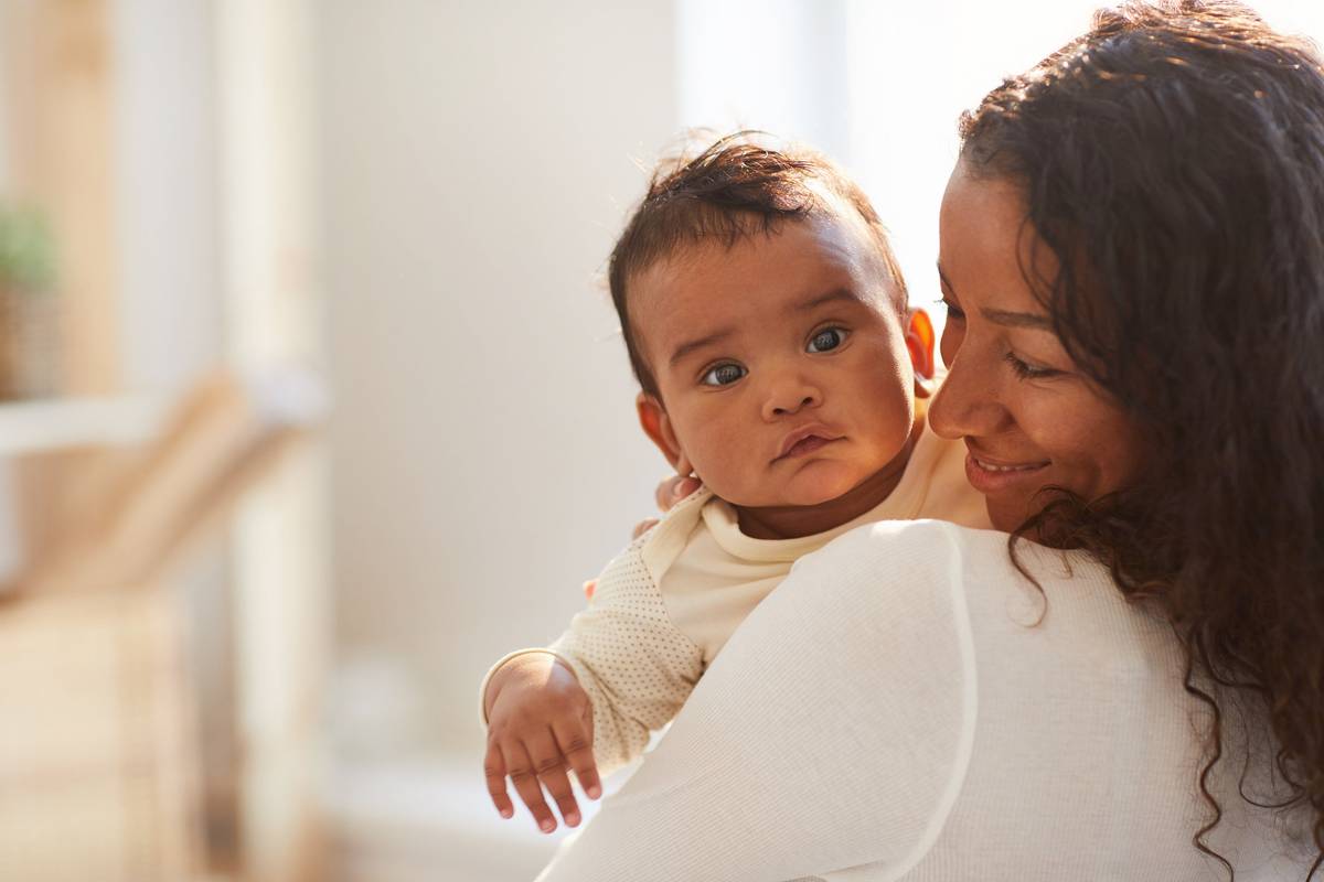 African mom with curly hair standing in room and holding adorable baby boy with chubby cheeks