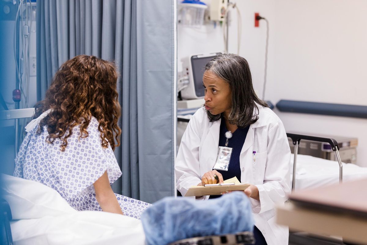 An unrecognizable teen girl wearing a hospital gown listens as the mature adult female emergency room doctor asks questions.