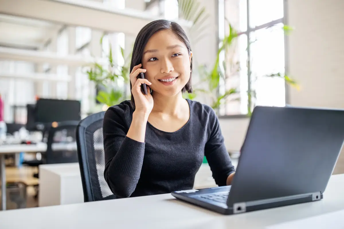 asian businesswoman sitting at her desk with laptop talking on mobile phone