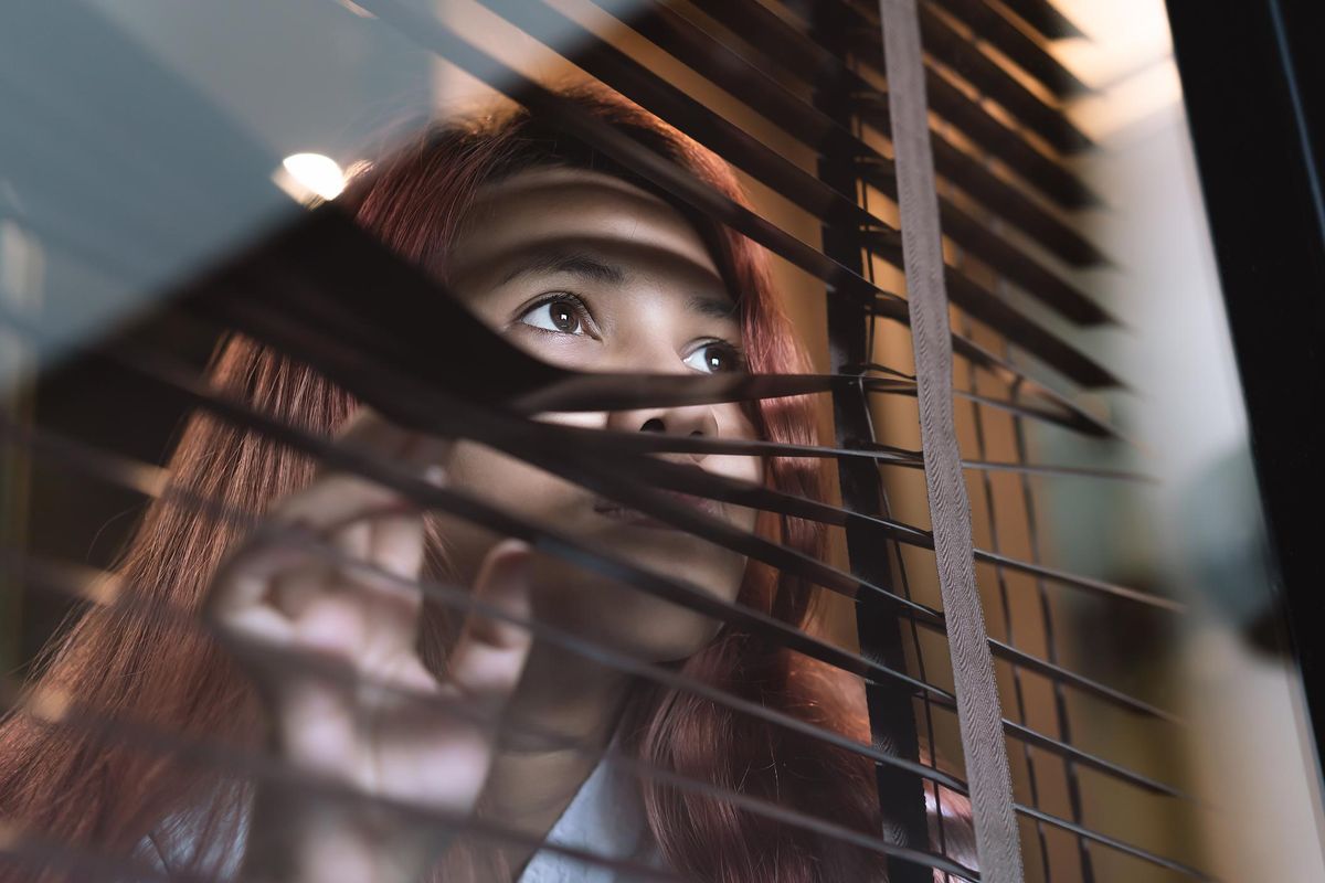 Asian diverse woman looking through window blinds