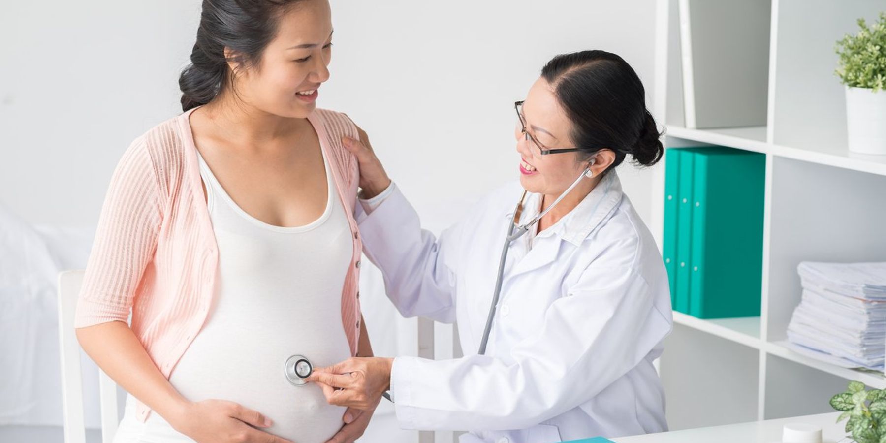 Asian doctor with stethoscope listening to belly of pregnant woman