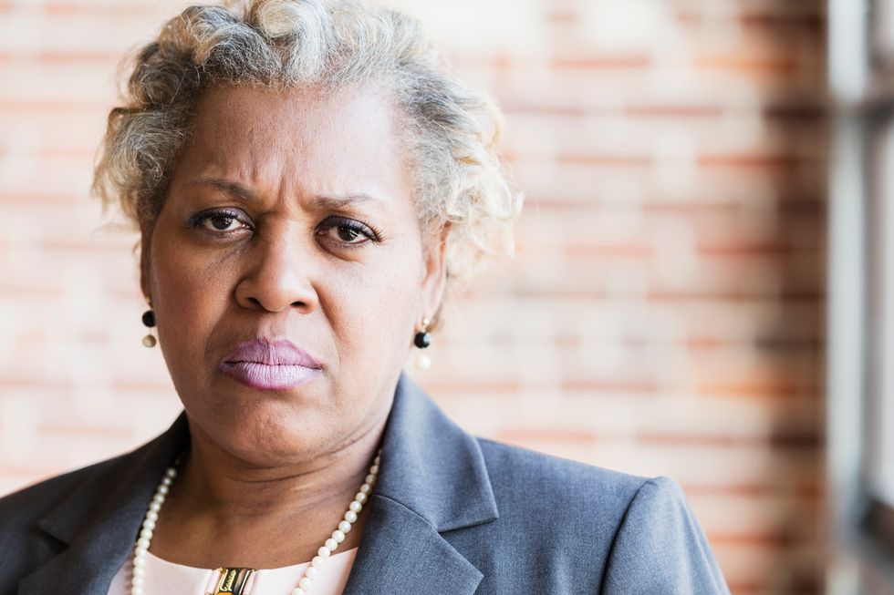 businesswoman wearing a jacket and pearl necklace. She has a serious, annoyed or angry expression on her face.