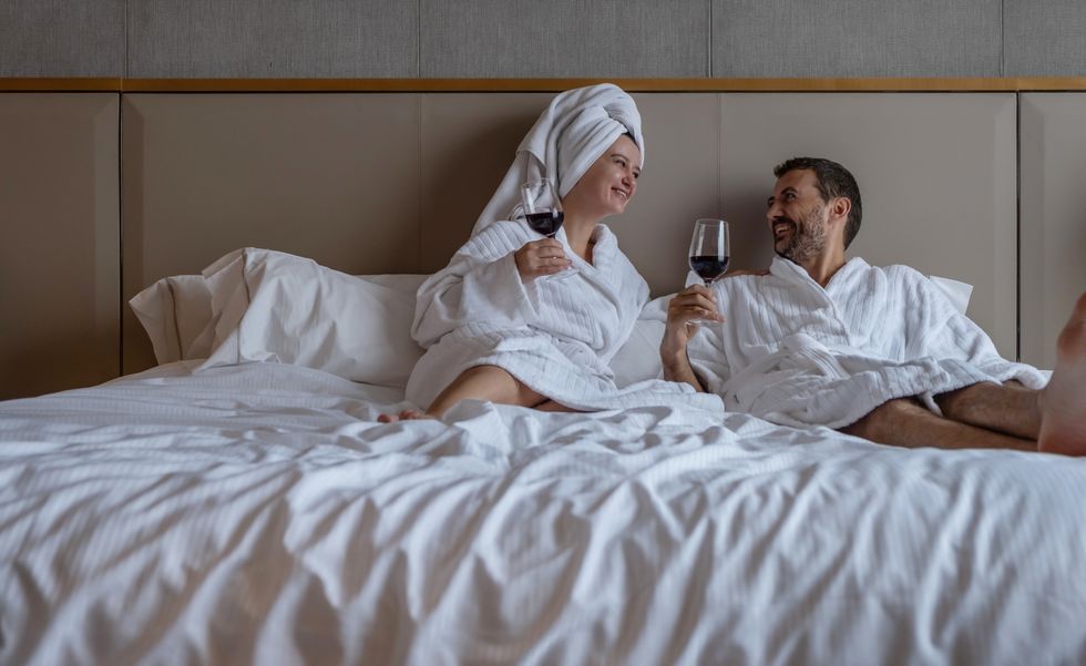 couple wearing white bathrobes smiling to each other and drinking wine in bed at hotel room