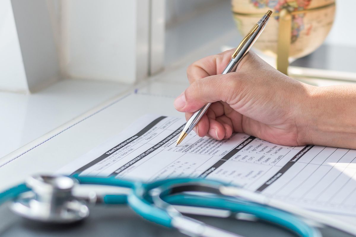 Doctor writing on medical health care record, patients discharge, or prescription form paperwork in hospital clinic office with physician's stethoscope on desk