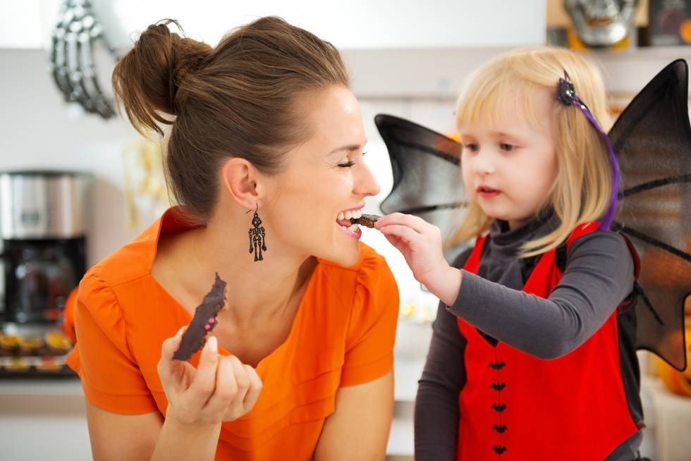 girl in bat costume with happy mother in decorated kitchen eating Halloween candy