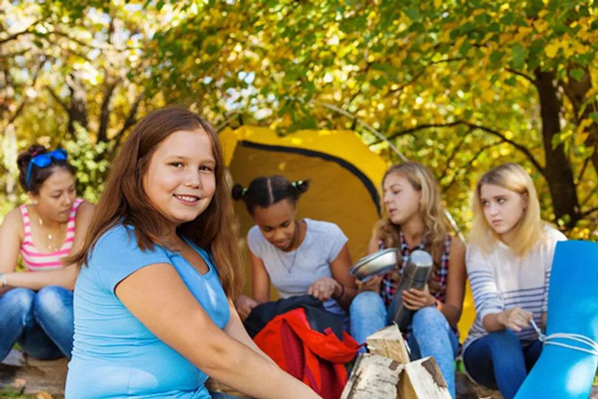 girl sitting near yellow tent with her friends during sunny autumn day at campsite