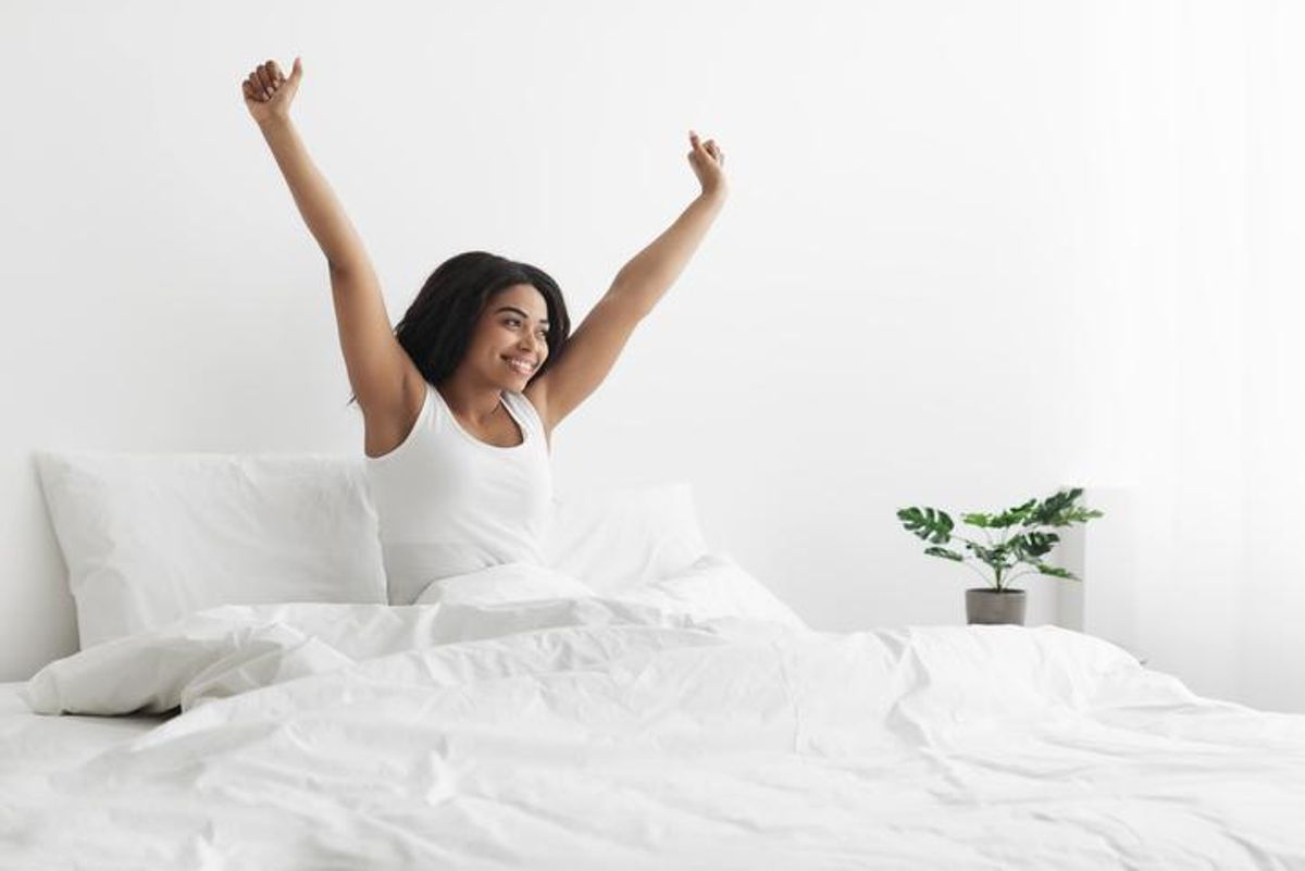 Happy african american woman sitting on bed with blanket, stretching after waking up