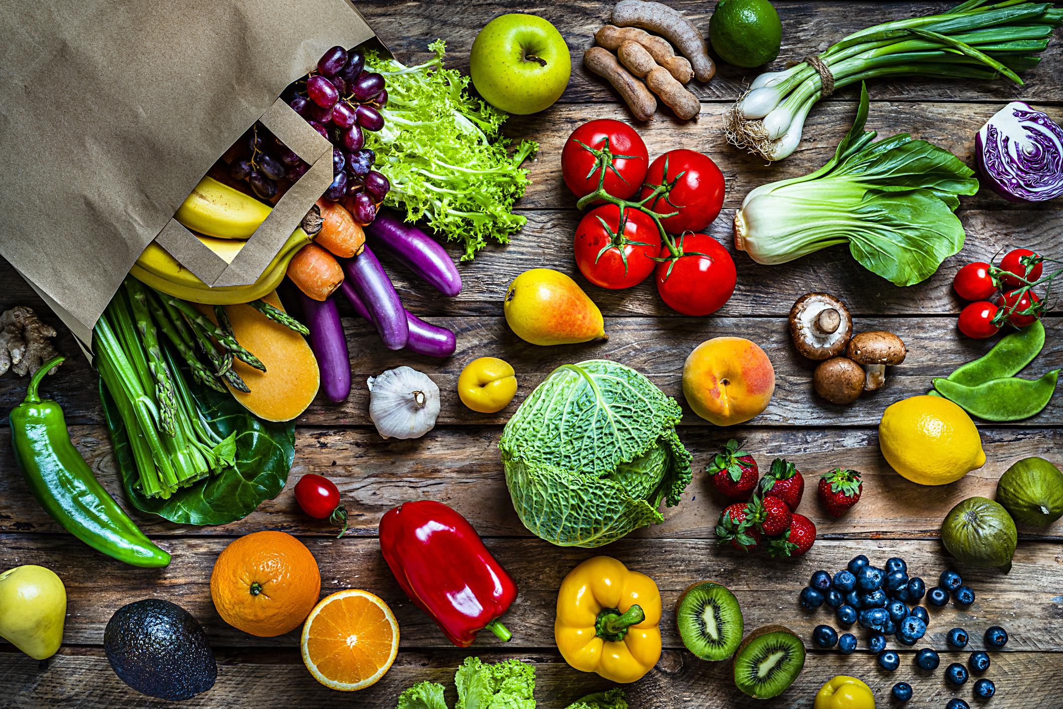 Healthy food: brown paper shopping bag filled with multicolored fresh organic fruits and vegetables shot from above on rustic wooden table. Fruits and vegetables included in the composition are apple, strawberry, banana, kiwi, orange, pear, grape, tamarind, blueberry, peach, lime, lemon, fig, kale, tomatoes, squash, asparagus, potato, celery, eggplant, carrots, lettuce, edible mushrooms, bell peppers, cauliflower, ginger, radish, avocado, onion, chili pepper, corn, among others
