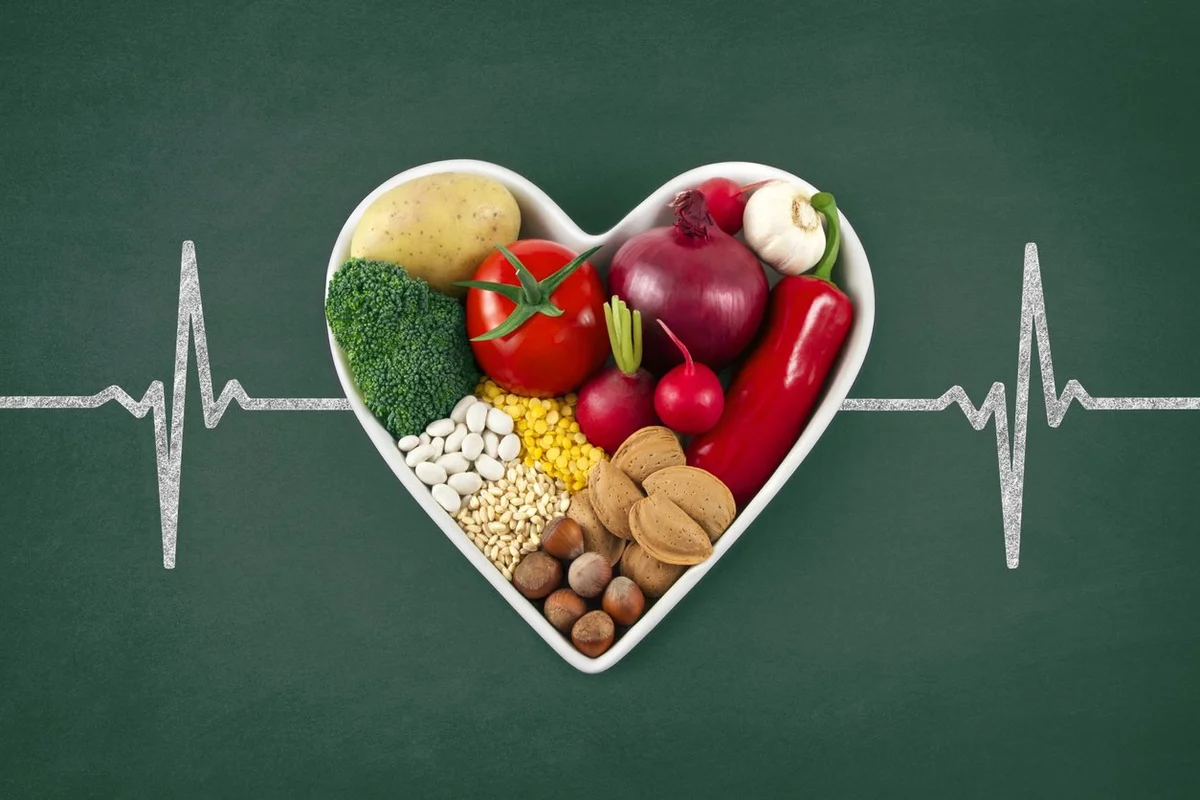 Heart health concept with related foods in white heart shaped bowl. potato red peppers, broccoli, radish, red onion, garlic, dry beans, almonds, nuts, and other legumes were arranged in heart shape plate on green blackboard with drawing of pulse trace.