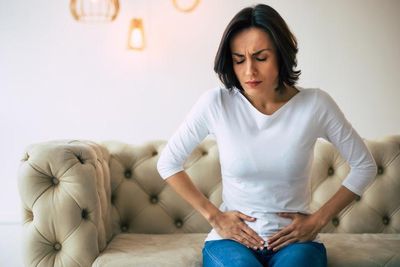 Adult woman is sitting on a sofa at home and touching her lower stomach while suffering from cramps