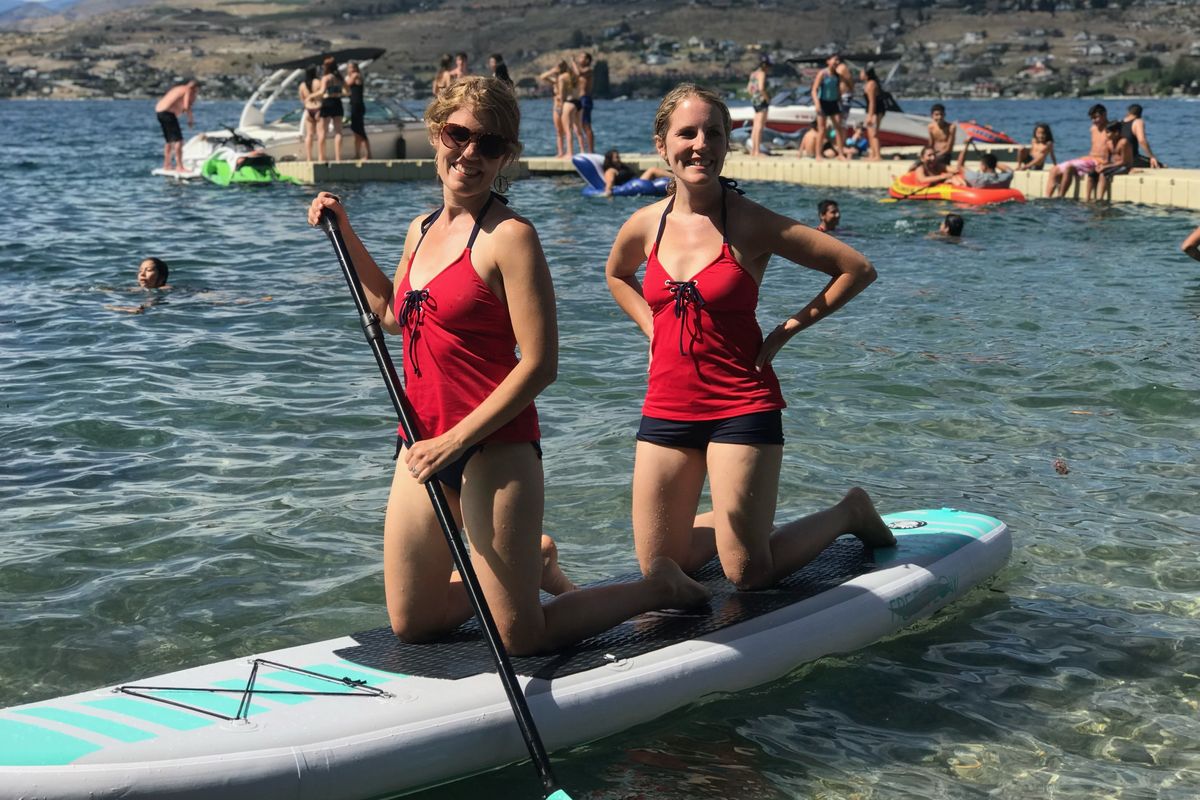 Joy and Jenelle are kneeling on a gray-and-teal paddleboard in the water. Jenelle has her hands on her hips and Joy has a paddle in her hands. Both sisters are wearing red tankini tops and black swimsuit bottoms; Joy is wearing sunglasses. 