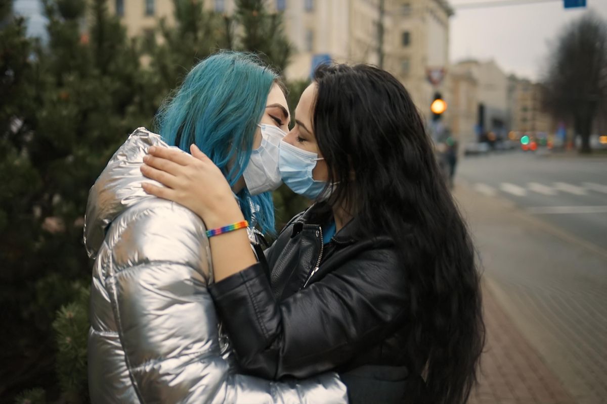 LGBT female couple kiss in medical face masks, one wears rainbow bracelet