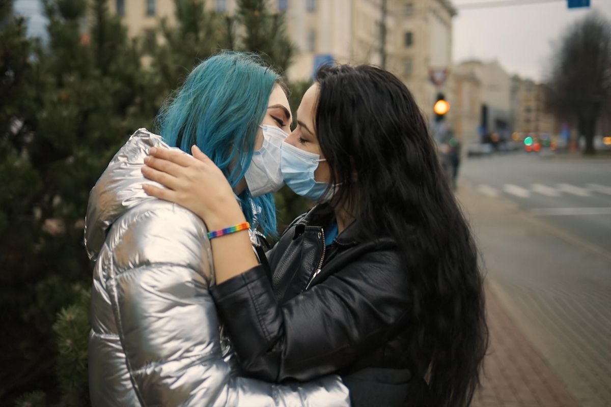 LGBT female couple kiss in medical face masks, one wears rainbow bracelet