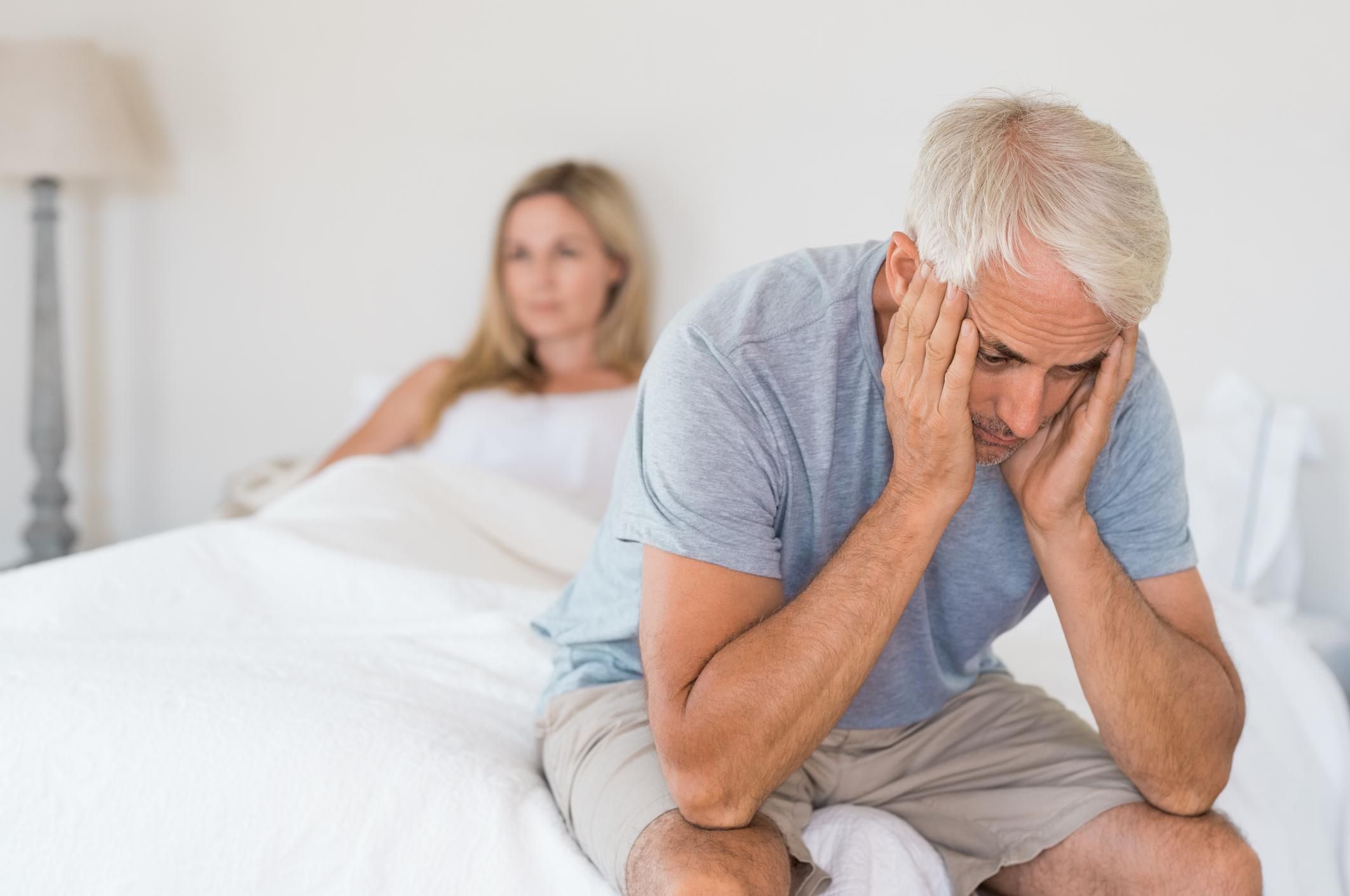 Man and woman in bed upset. Man sitting on the edge of the bed holding head in hands.