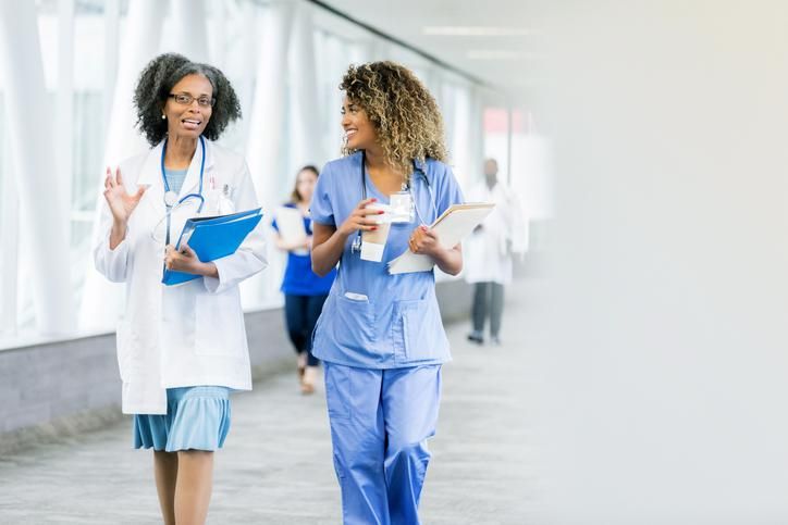 Mature female doctor gestures while walking in hospital skybridge with female nurse