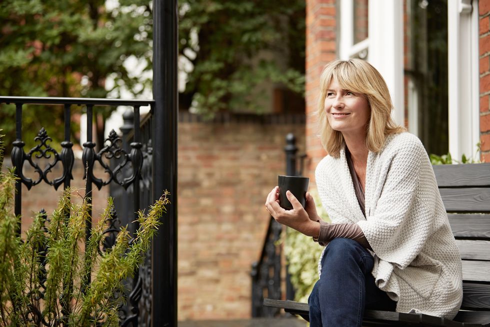 mature woman sitting on a bench on her front porch drinking a coffee