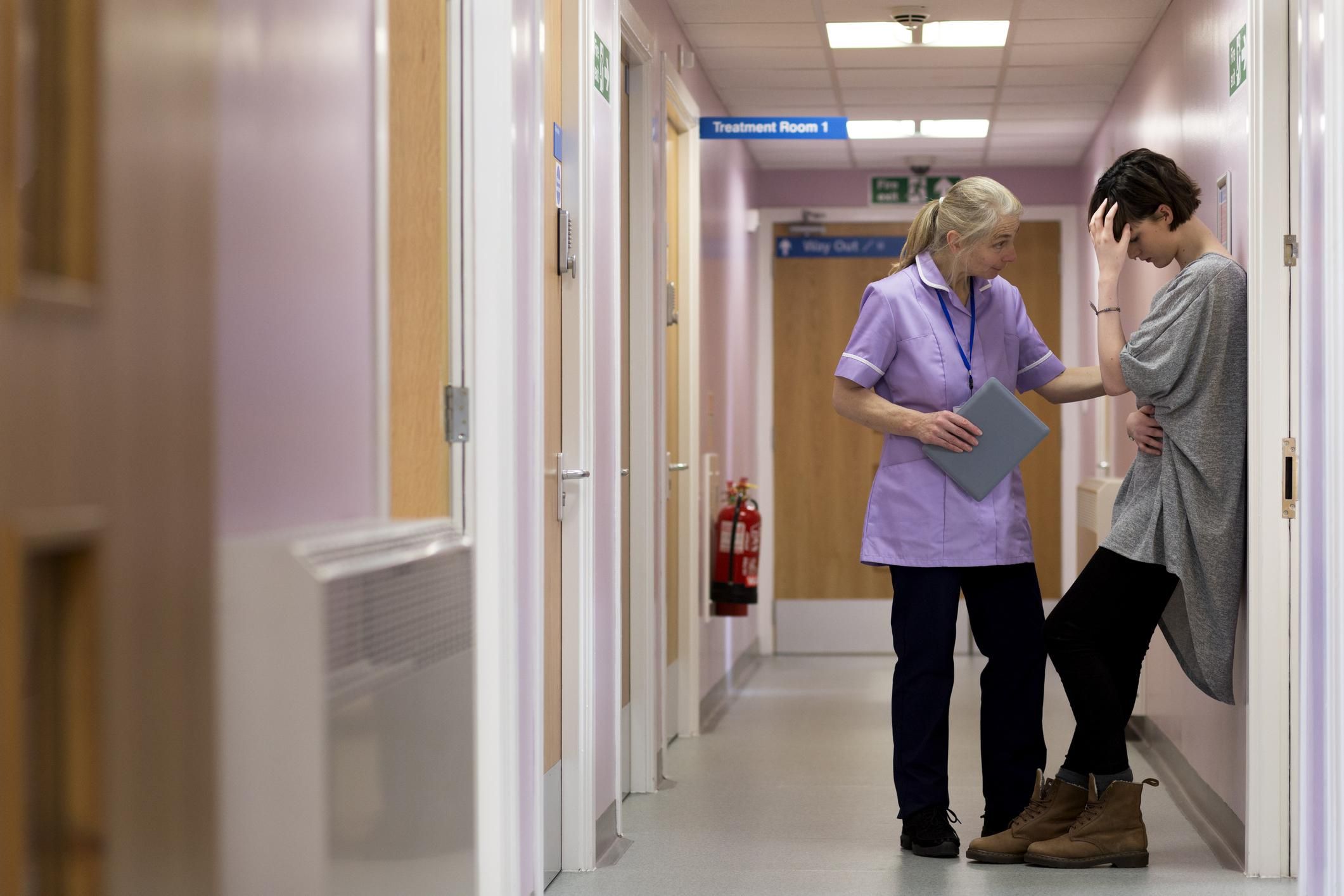 Medical woman comforts young female adult as she stands with her back against to the wall in a hospital corridor.
