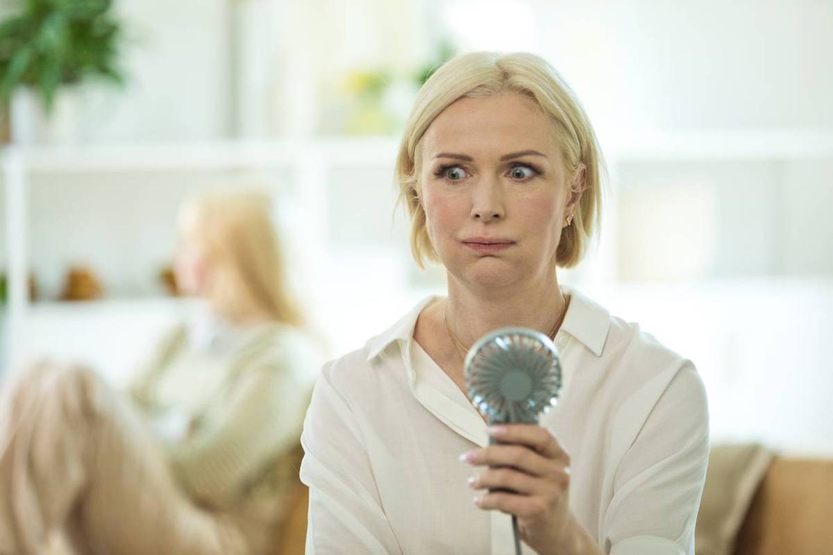 Menopausal mature woman sitting on sofa in the living room at home having a hot flash and using fan