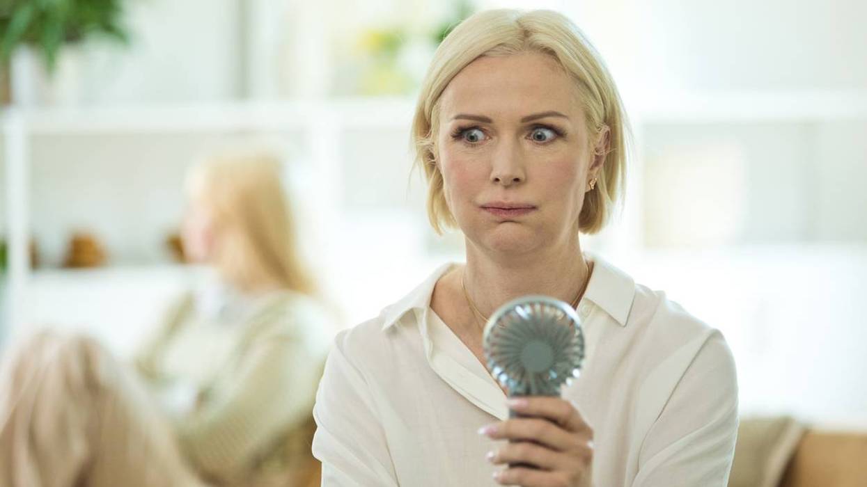 Menopausal mature woman sitting on sofa in the living room at home having a hot flash and using fan