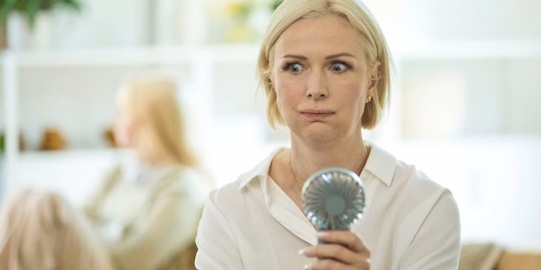 Menopausal mature woman sitting on sofa in the living room at home having a hot flash and using fan