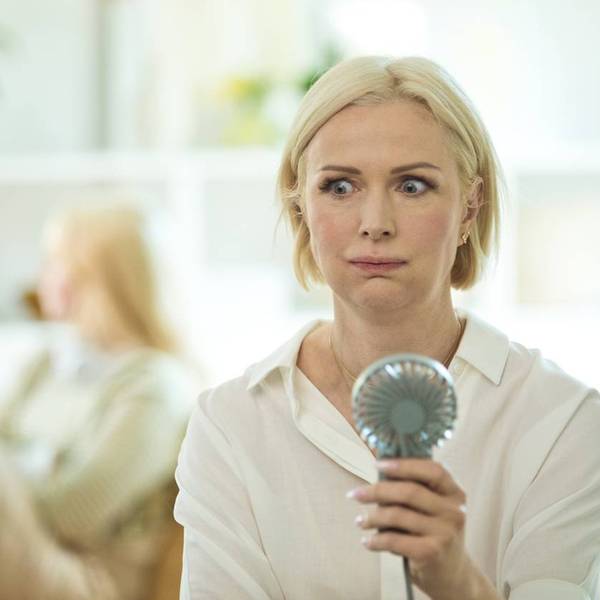 Menopausal mature woman sitting on sofa in the living room at home having a hot flash and using fan