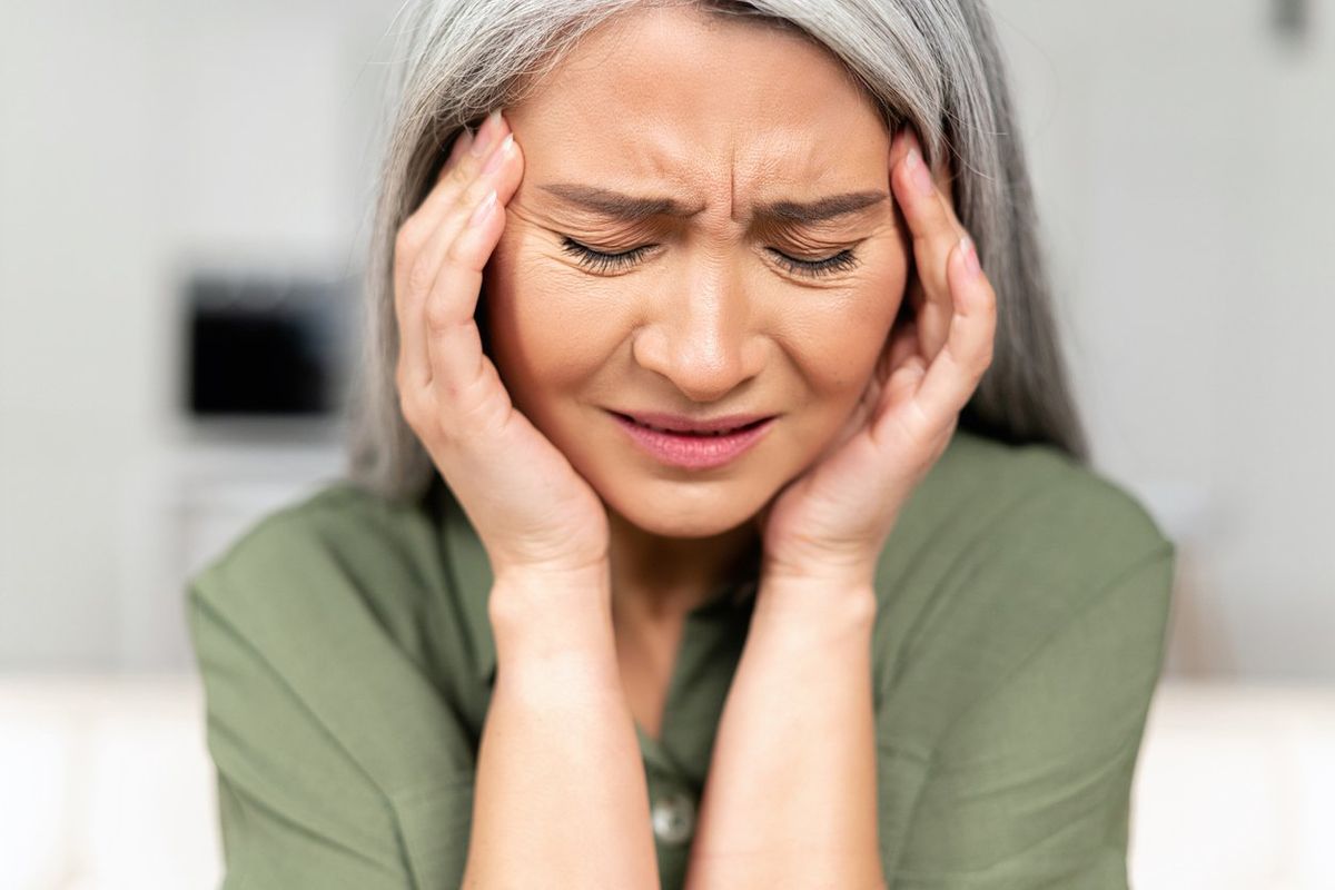 Middle-aged woman with long silver hair rubbing temples, suffering from migraine