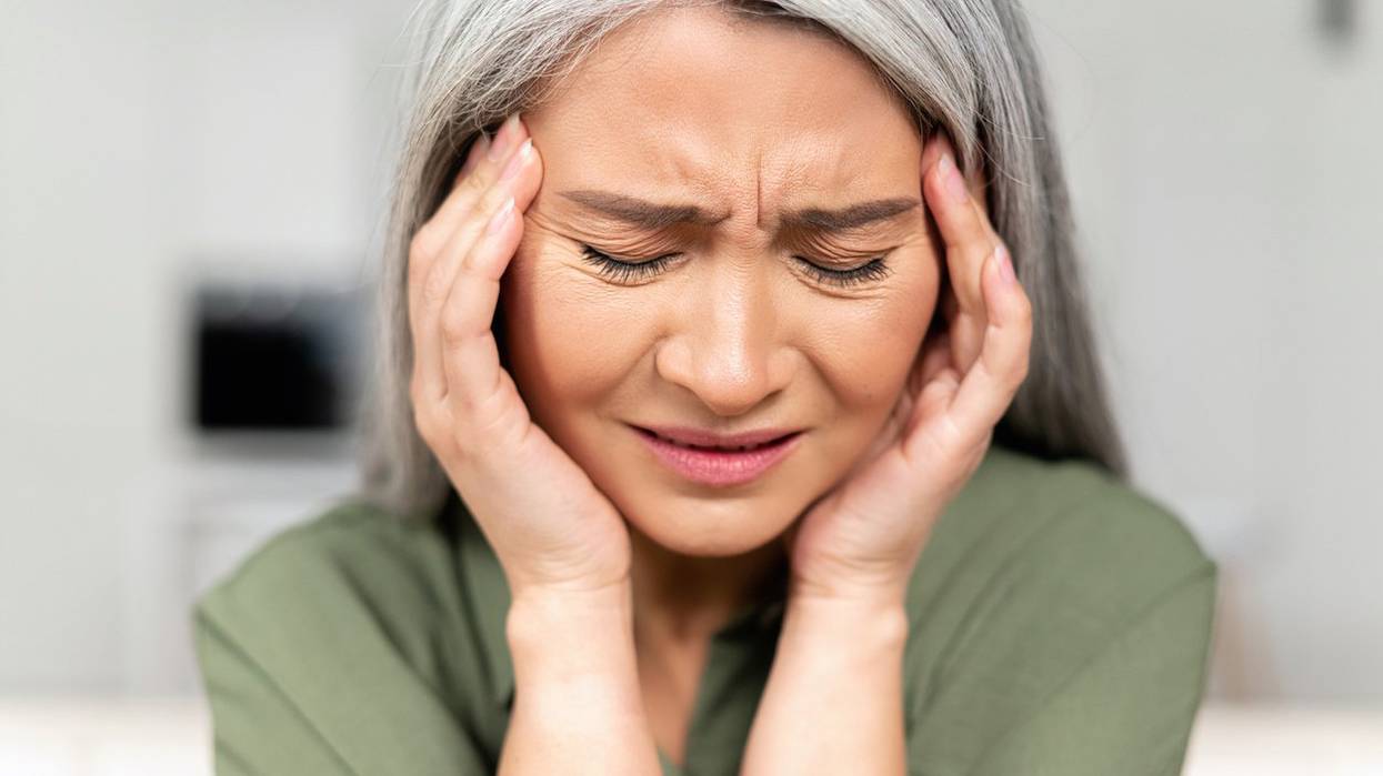 Middle-aged woman with long silver hair rubbing temples, suffering from migraine