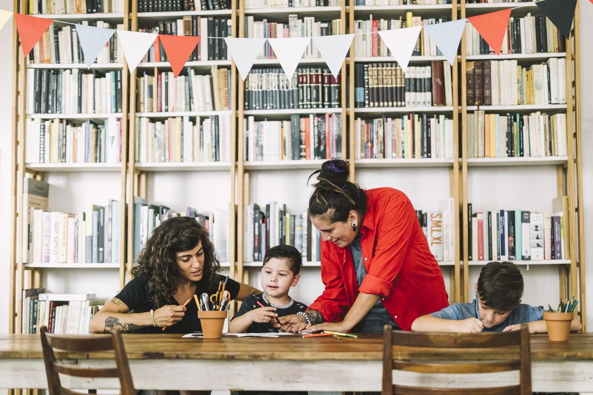 Mothers assisting son in drawing at table. Homosexual parents and children are against bookshelf. Women are with boys at home.
