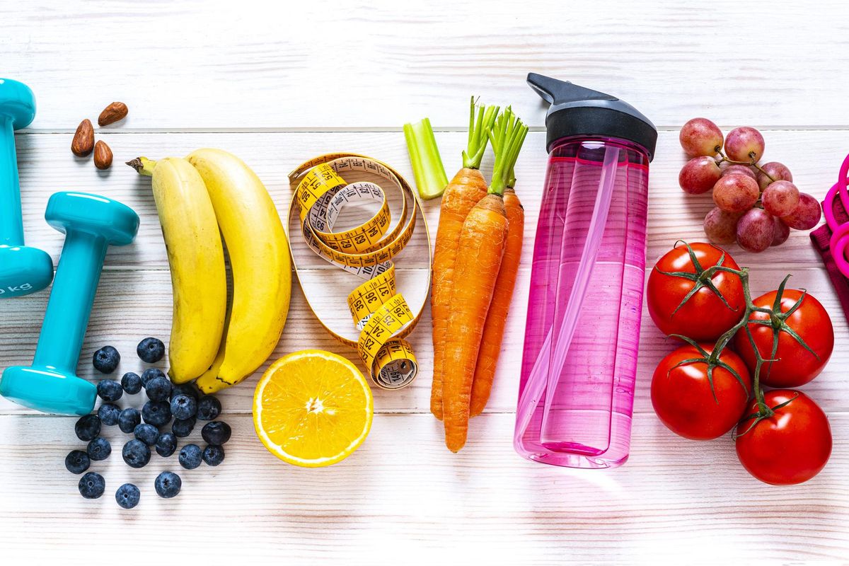 rainbow colored dumbbells, jump rope, water bottle, towel, tape measure and healthy fresh organic vegetables, fruits and nuts arranged side by side