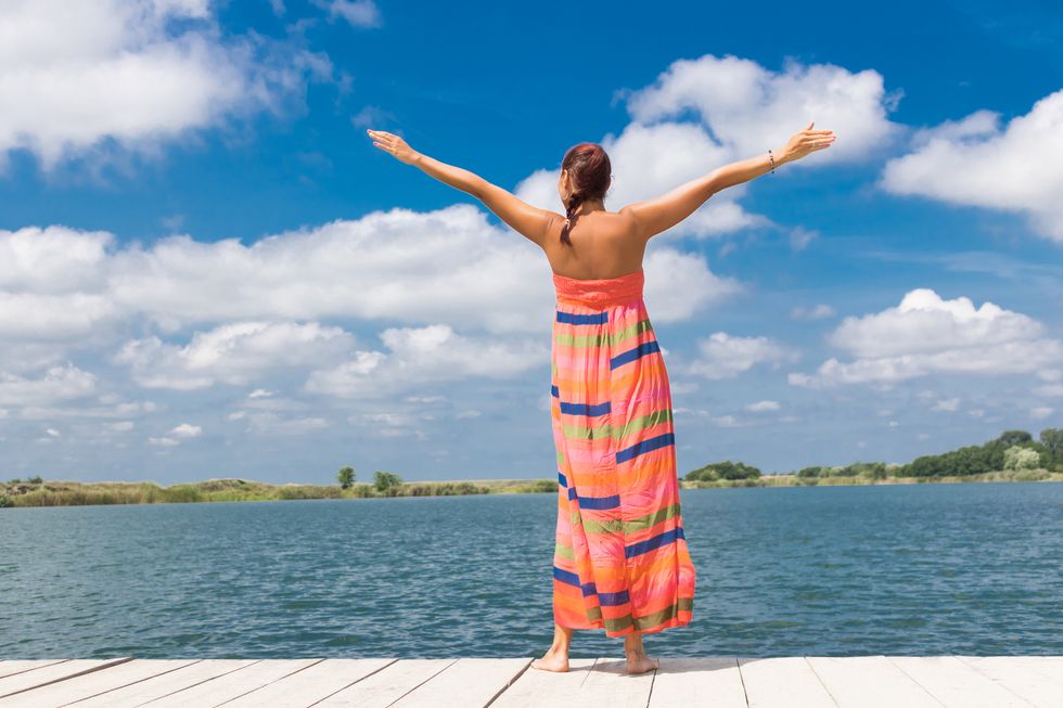 Rear view of carefree woman with arms outstretched standing on a pier and enjoying in summer freedom by the water.