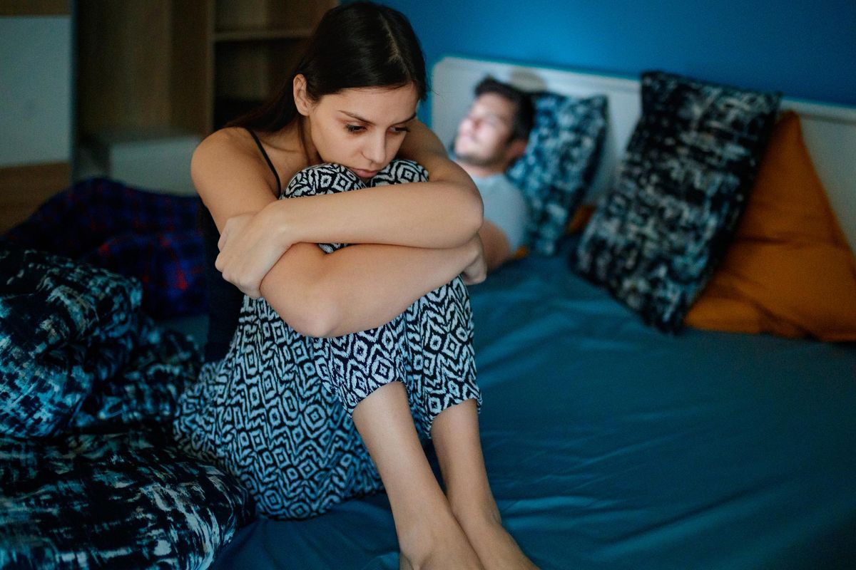 Sad woman sitting on bed with partner in background