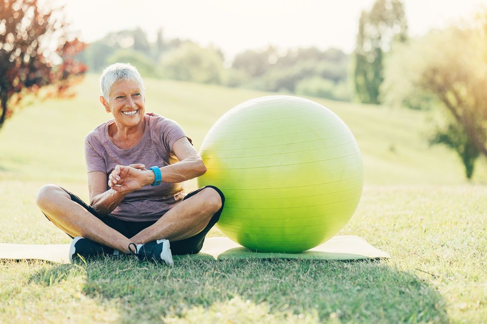 Senior sportswoman with smart watch and fitness ball sitting on the grass