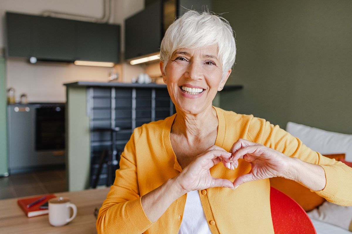 Senior woman is at home, she is in the living room and showing a heart-shaped symbol