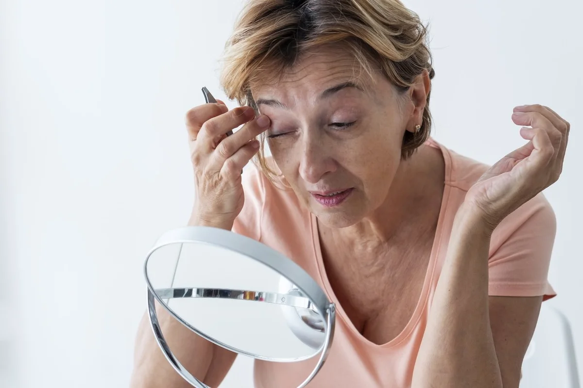 Senior woman shaping her eyebrows with tweezer at home.