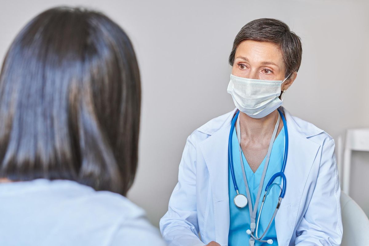 Serious middle-aged woman general practitioner GP in face mask consulting female patient during covid-19 outbreak, doctor giving recommendations and explaining medical diagnosis to client in clinic
