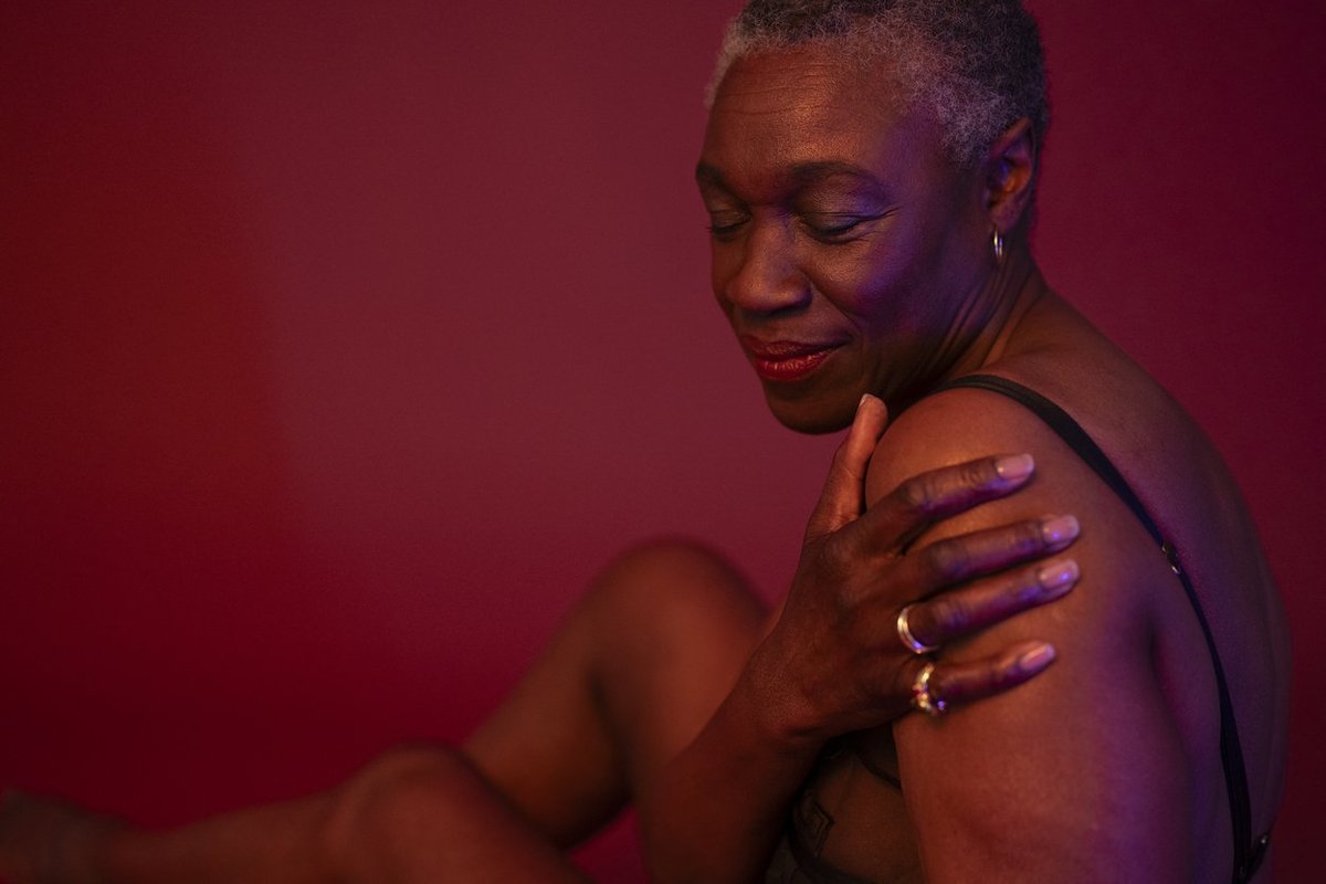 shorthaired woman in semi-dress seated against rose colored background, hand on upper arm, eyes closed