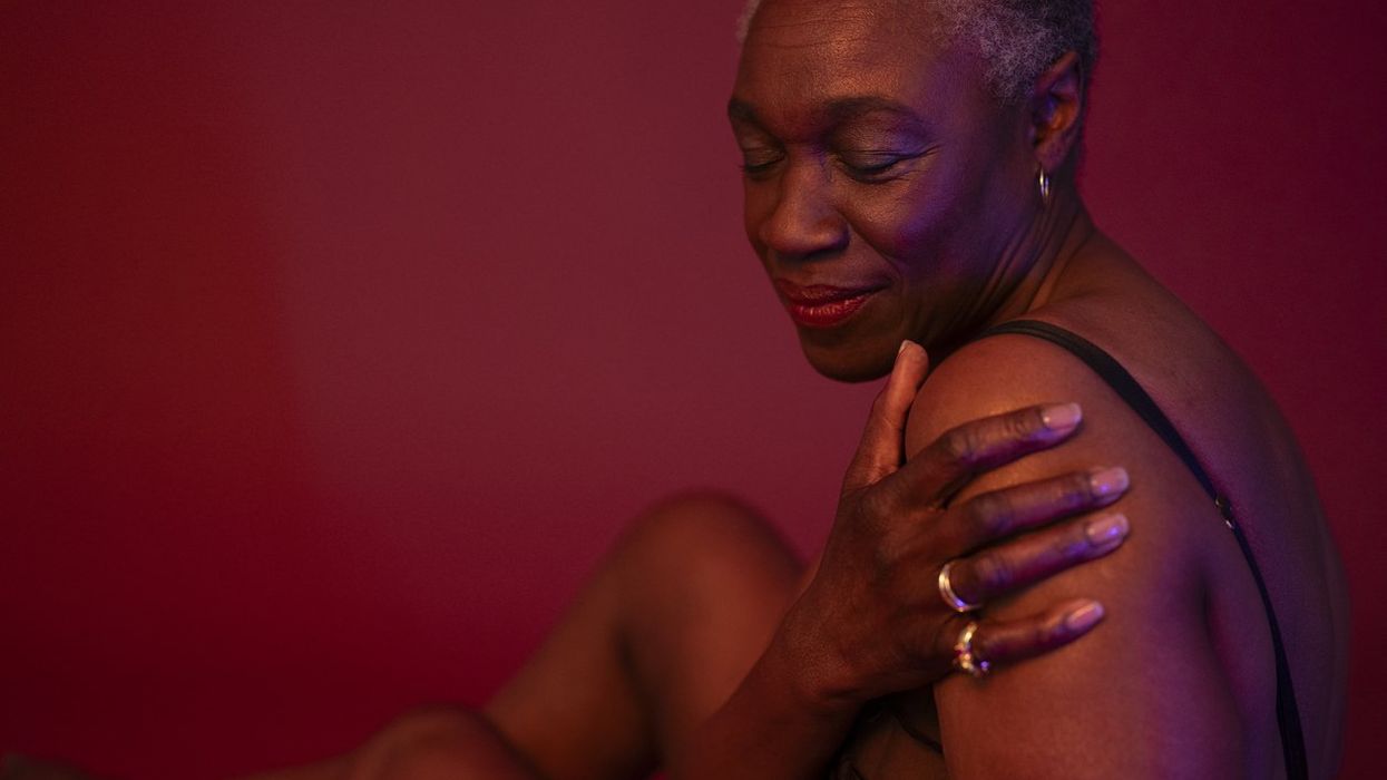 shorthaired woman in semi-dress seated against rose colored background, hand on upper arm, eyes closed