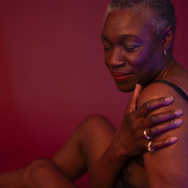 shorthaired woman in semi-dress seated against rose colored background, hand on upper arm, eyes closed