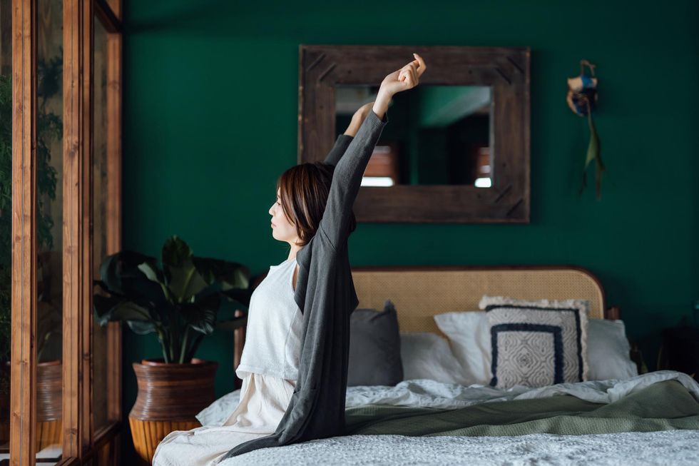 Side profile of young Asian woman sitting in bed, stretching arms after waking up in the morning against sunlight.