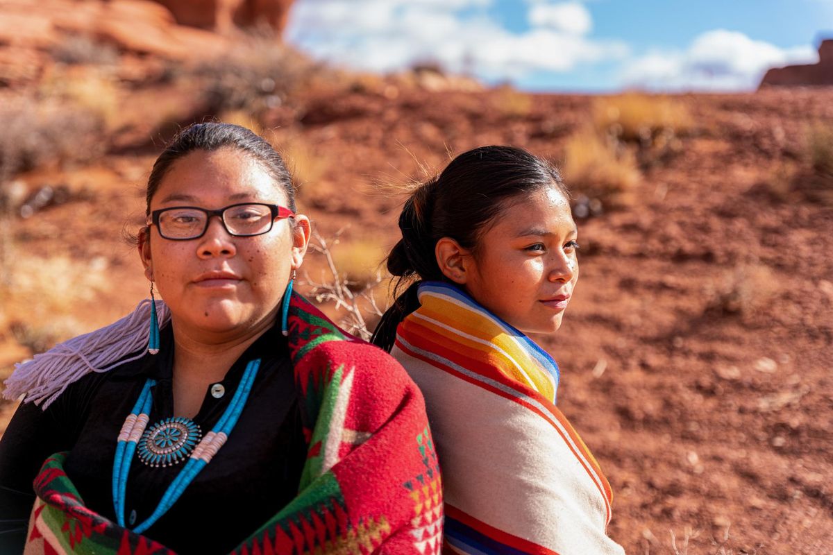 Two Native American, Navajo Sisters Dressed In Traditional Clothing, Wrapped In A Navajo Blanket Sitting Back To Back, One Looking Forward, The Other Looking Away